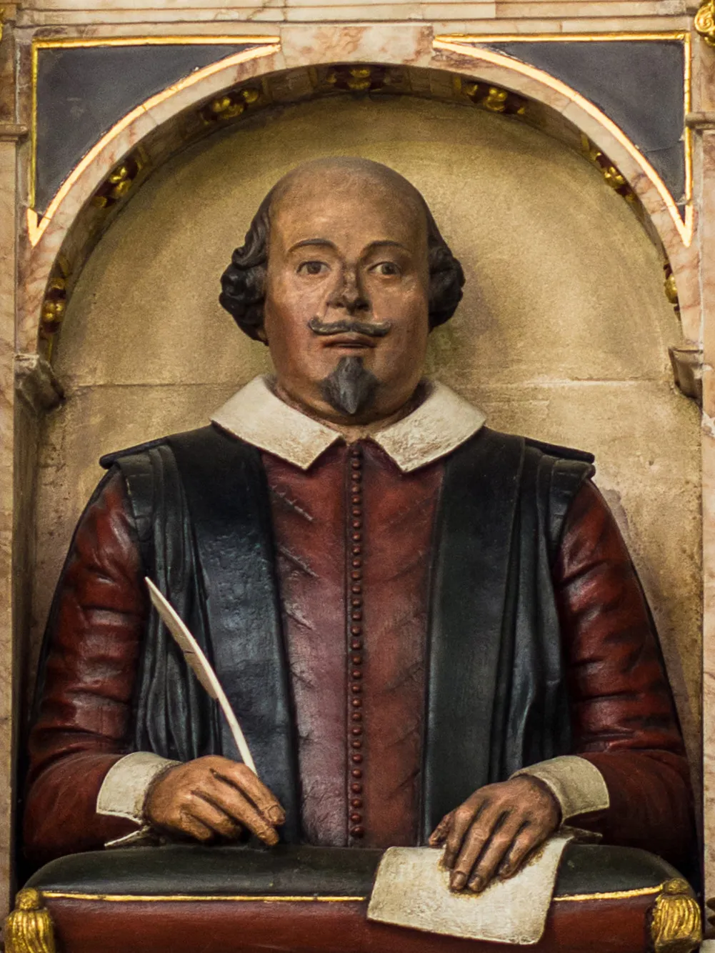 A painted limestone carving of the upper half of Shakespeare, who is bald with a fringe of hair about the ears. He is wearing a red shirt, a black vest, and a white collar. He is holding a quill pen in his right hand and a sheet of paper in his left hand.
