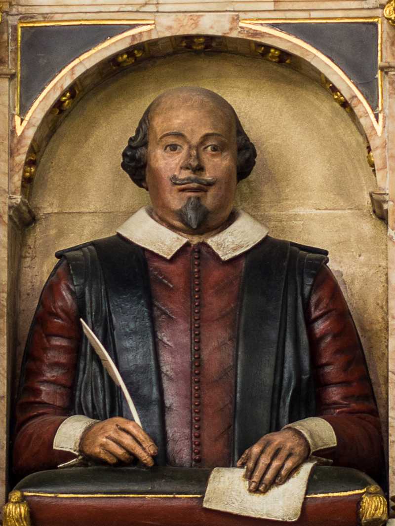Image of a painted limestone carving of the upper half of Shakespeare, who is bald with a fringe of hair about the ears. He is wearing a red shirt, a black vest, and a white collar. He is holding a quill pen in his right hand and a sheet of paper in his left hand.